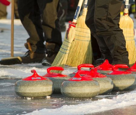 Grey curling stones lined up on the ice. Behind them there are three brooms being held by people wearing black snow pants (no faces are visible)  